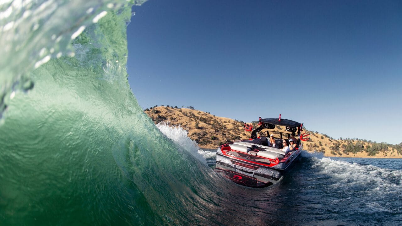 A speedboat with passengers navigates through a wave on a sunny day with hills in the background.