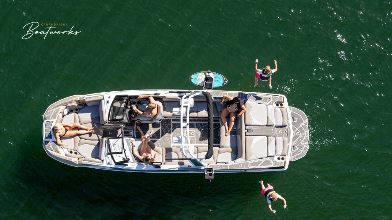 Aerial view of a boat on the water with four people. Two are seated inside, one is standing at the rear, and one person is jumping into the water.