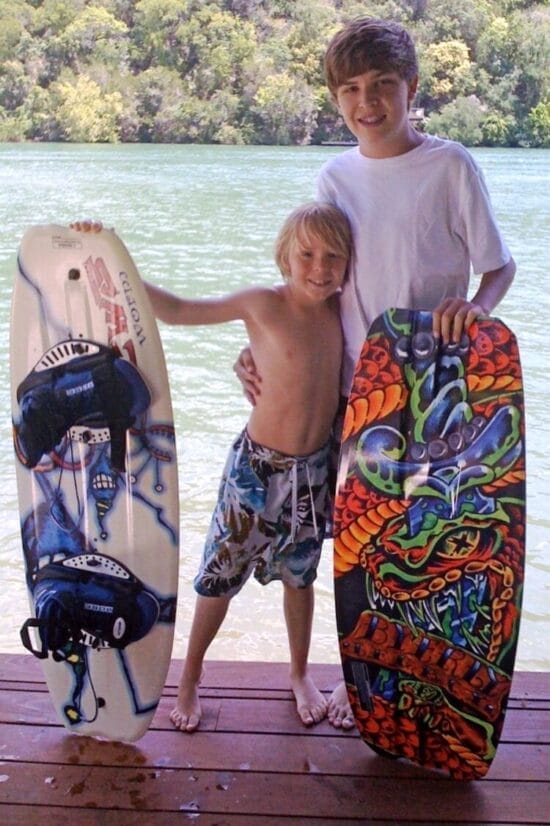 Two kids are standing on a dock by a lake, each holding a wakeboard. The younger child is shirtless, and the older child is wearing a white shirt. Trees are visible across the water.