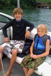 Two kids sitting on a boat. The older one is wearing a black shirt and patterned shorts, while the younger one is in a life vest and floral shorts. Water and a wooden house are in the background.