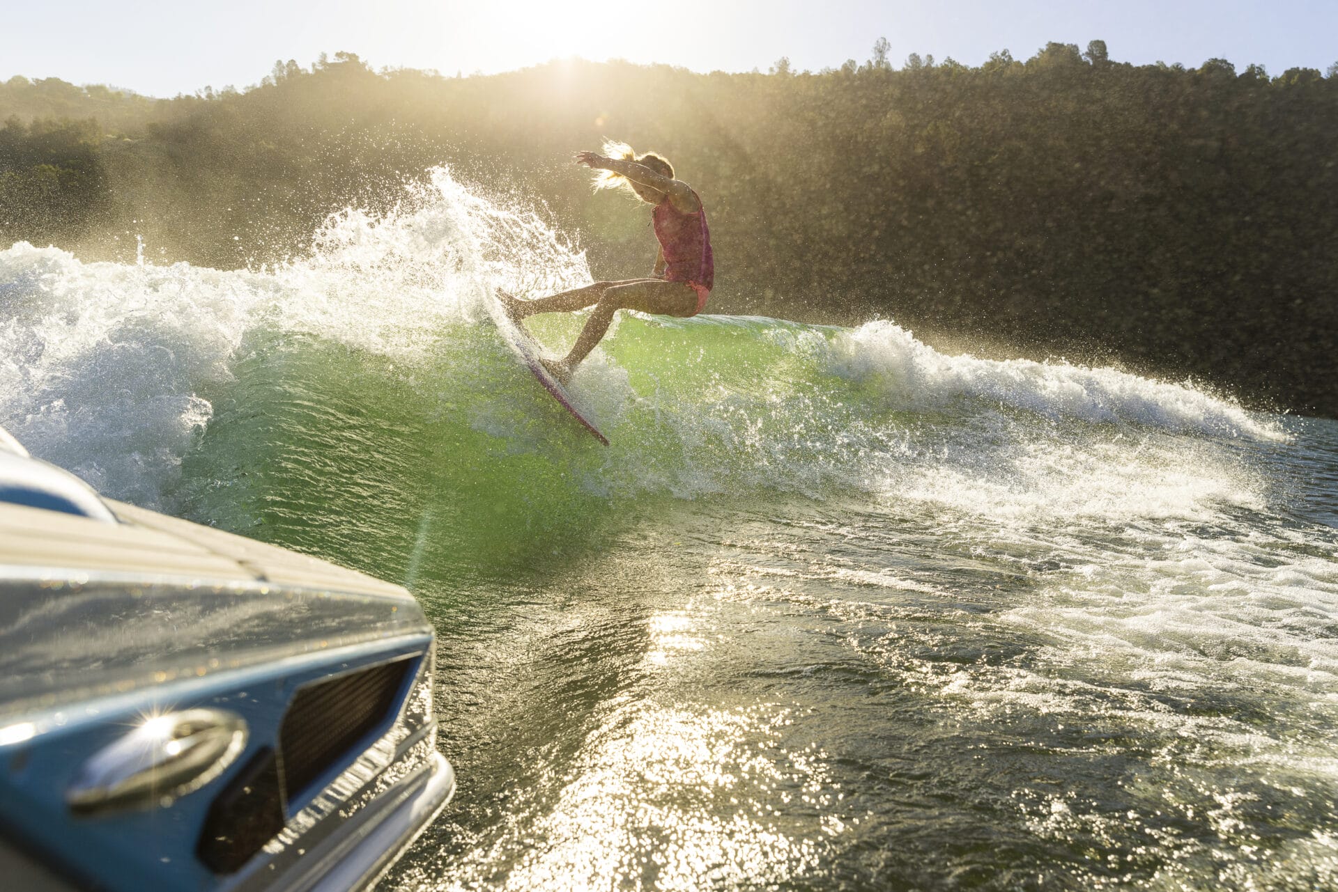 A person surfs on a wave behind a boat in a sunny outdoor setting with a forested hillside in the background.