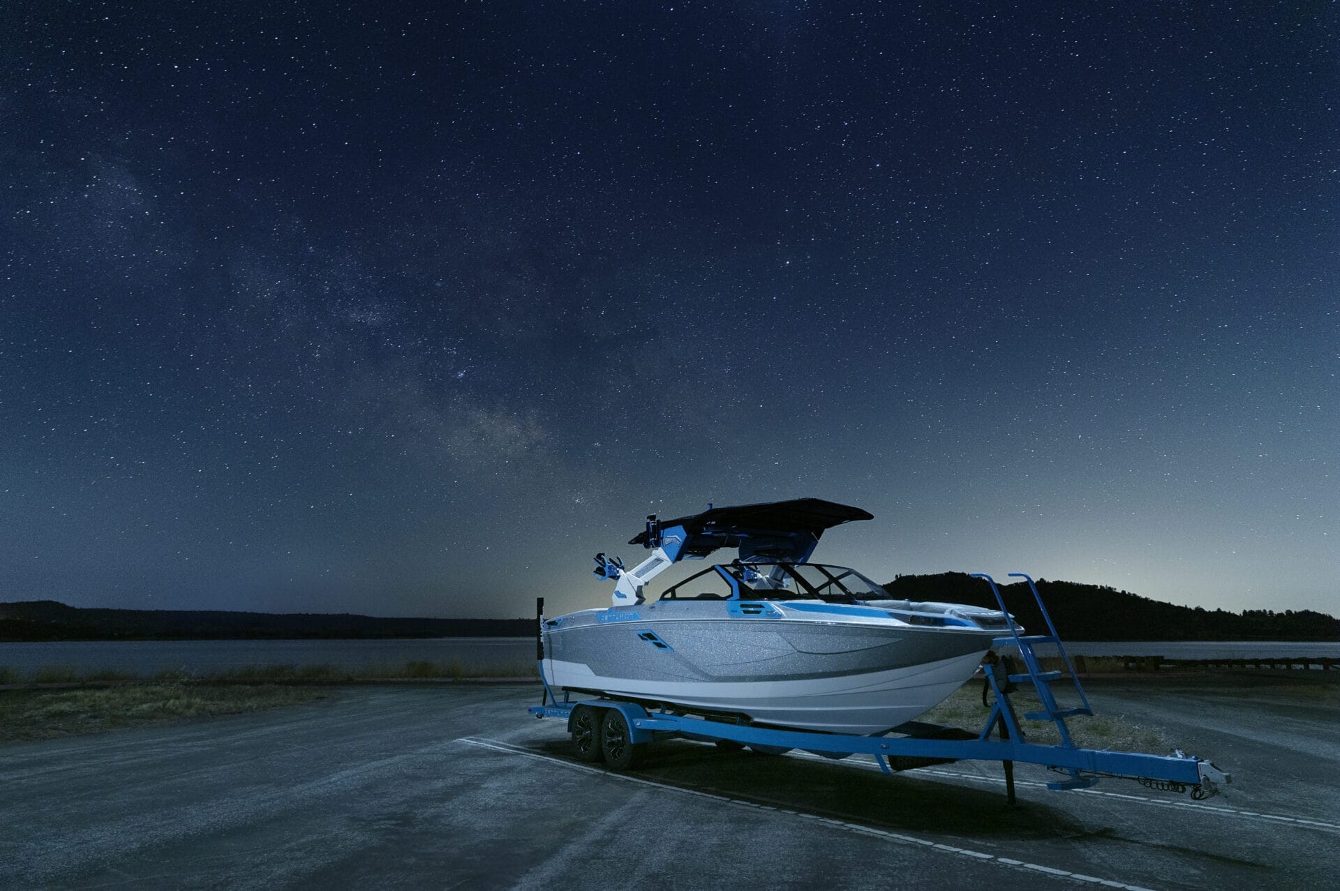 A boat on a trailer is parked on a lakeside road at night, with a starry sky in the background.