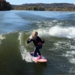 A person in a wetsuit rides a wave on a pink surfboard, making hand gestures, with a scenic view of trees and hills in the background.