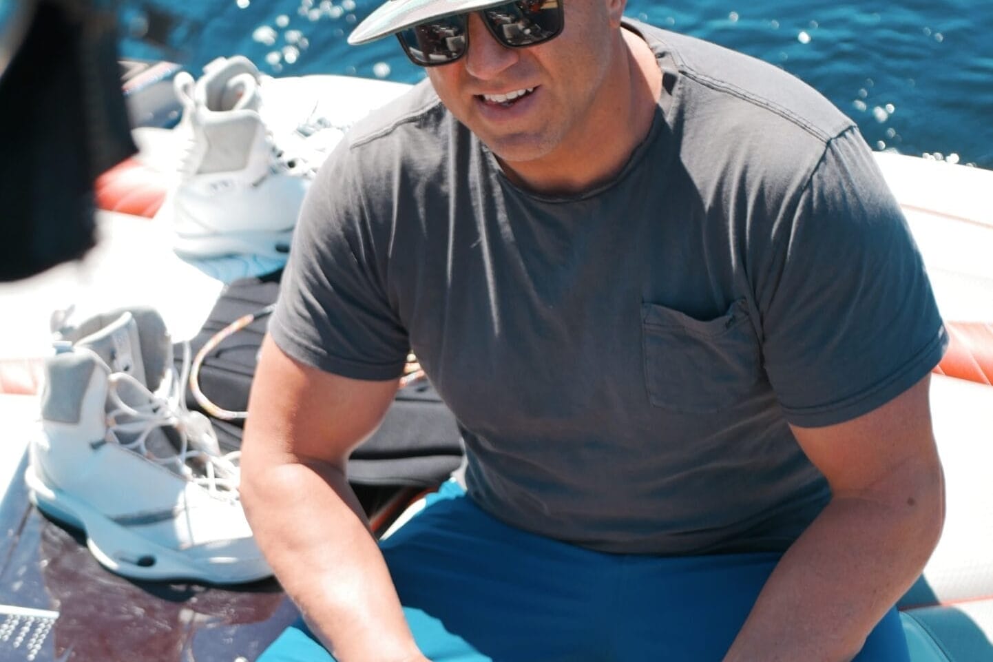 A man wearing a camouflage hat, sunglasses, grey t-shirt, and blue shorts sits on a boat near water sports gear with water in the background.