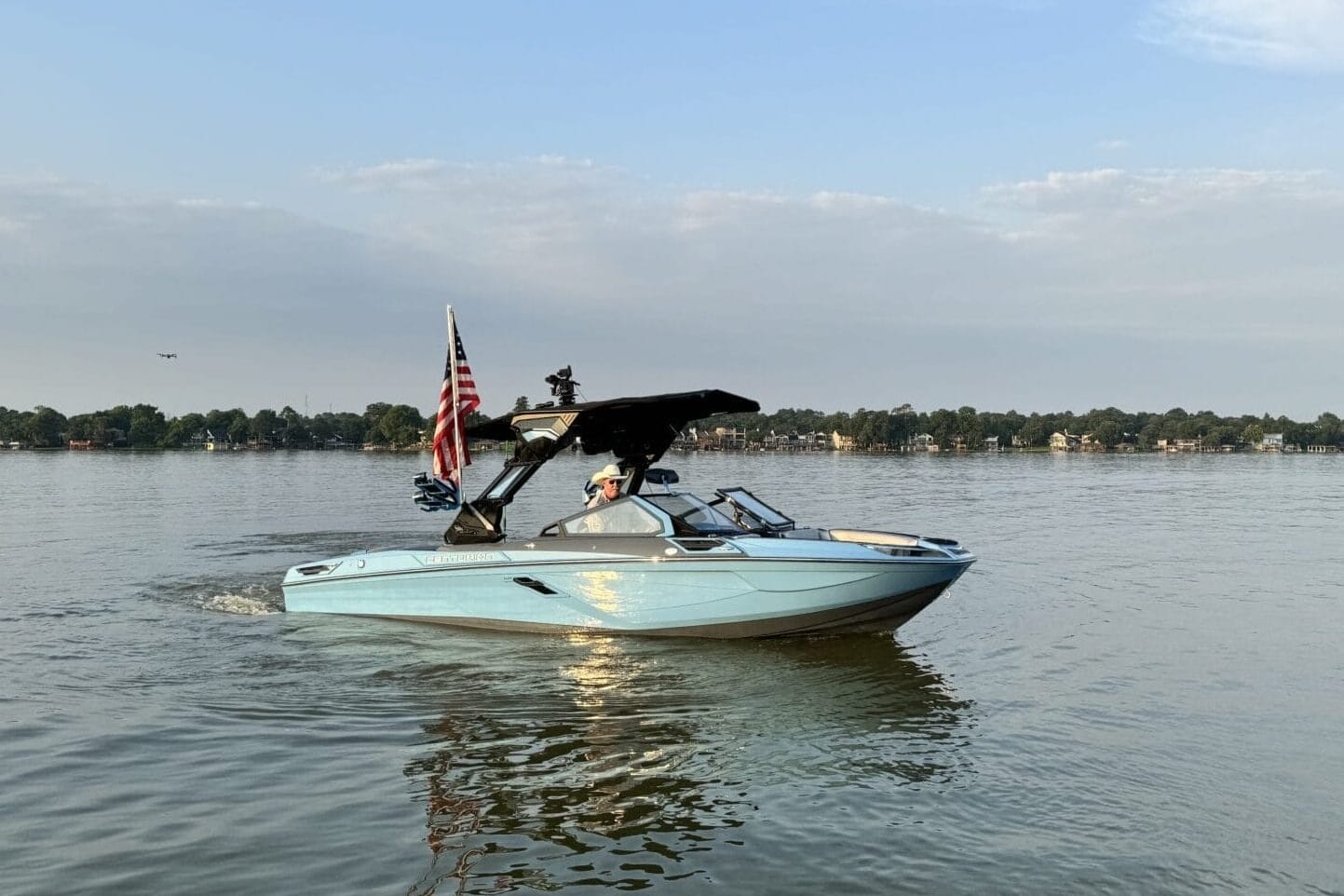 A light blue motorboat with an American flag moves across a calm lake under a partly cloudy sky, with a tree-lined shore in the background.
