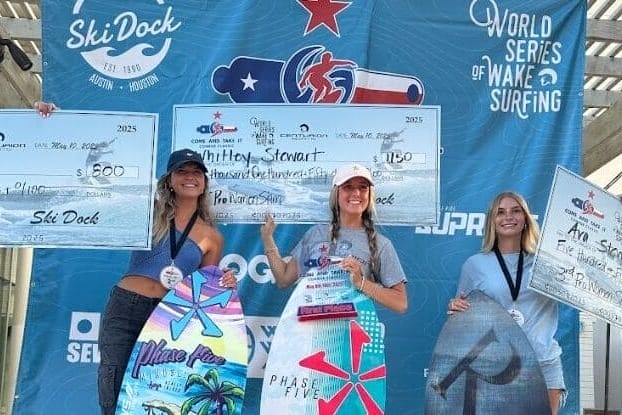 Three women stand on a podium holding wakeboards and oversized prize checks at a wake surfing competition, with event banners in the background.