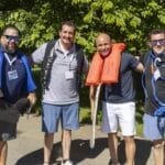 Four men standing outdoors wearing casual clothes, each holding or wearing boating safety gear, including life jackets, rope, and a paddle, with trees in the background.