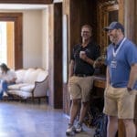 Two men stand and talk in a wooden hallway while a woman sits on a couch in the background using her phone.