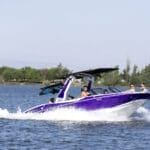 A group of people ride a purple and white motorboat on a calm lake with trees and clear skies in the background.