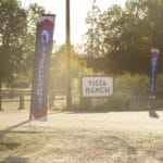 Two vertical flags and a sign reading "Vista Ranch" are visible along a sunlit rural road with trees in the background.