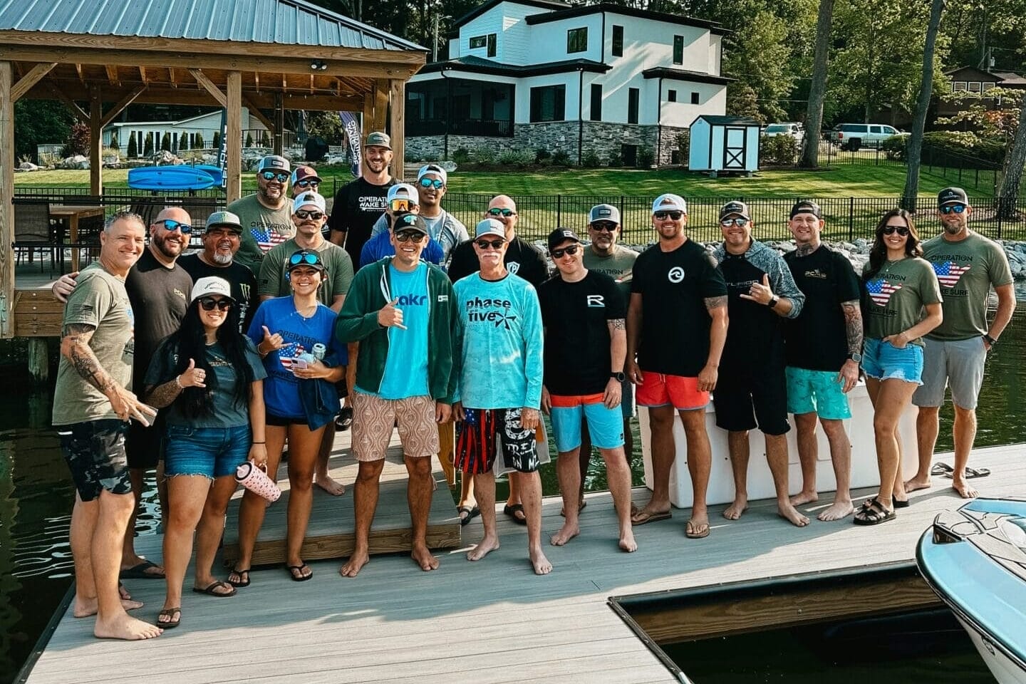 A group of people pose for a photo on a dock by the water, with trees, a gazebo, and a house visible in the background.