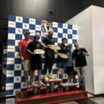 Six men pose on a winners' podium in front of a checkered backdrop, holding award plaques, with one man standing on the tallest platform.