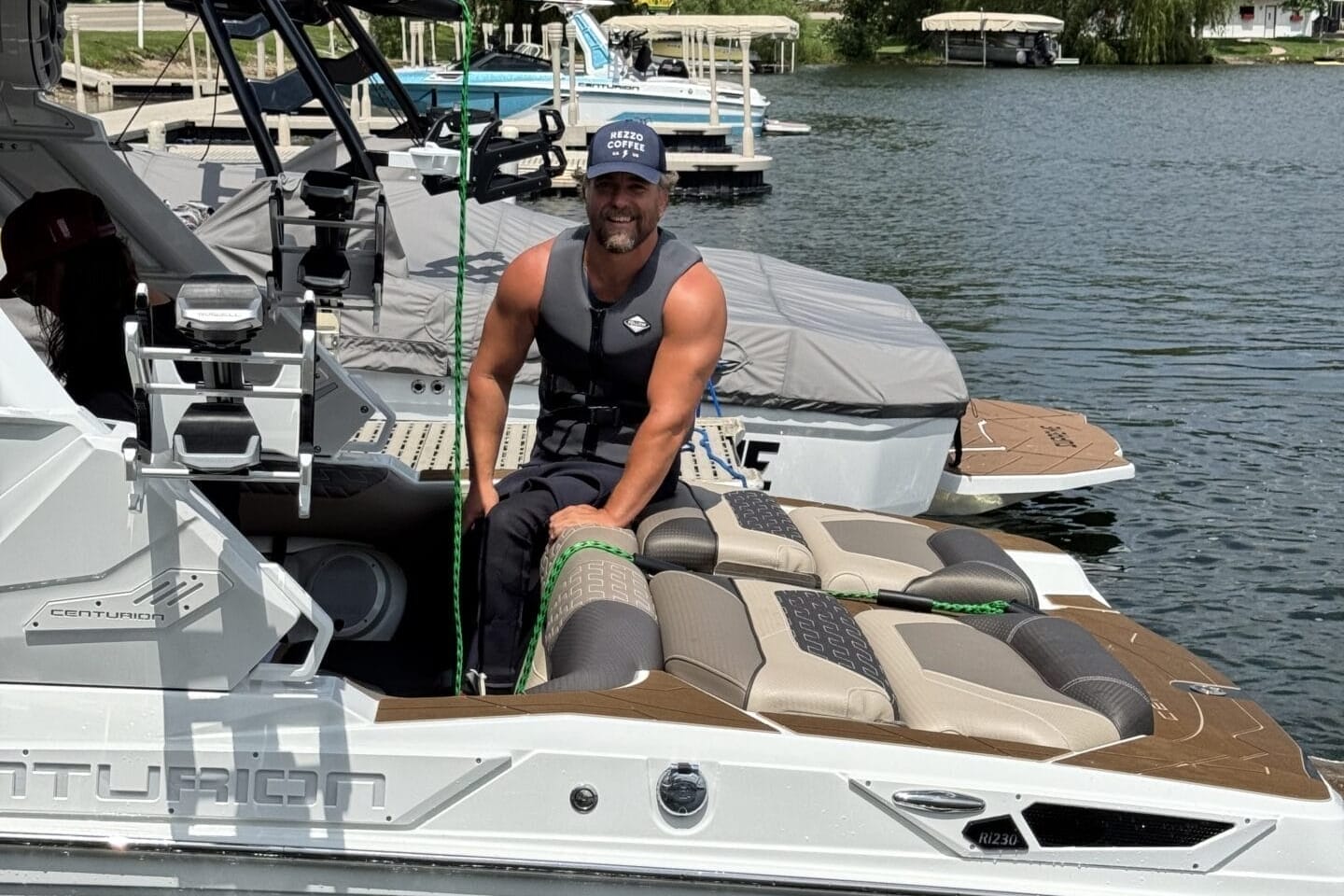 A person wearing a life jacket and cap sits on the edge of a boat docked on a lake, surrounded by trees and other boats.