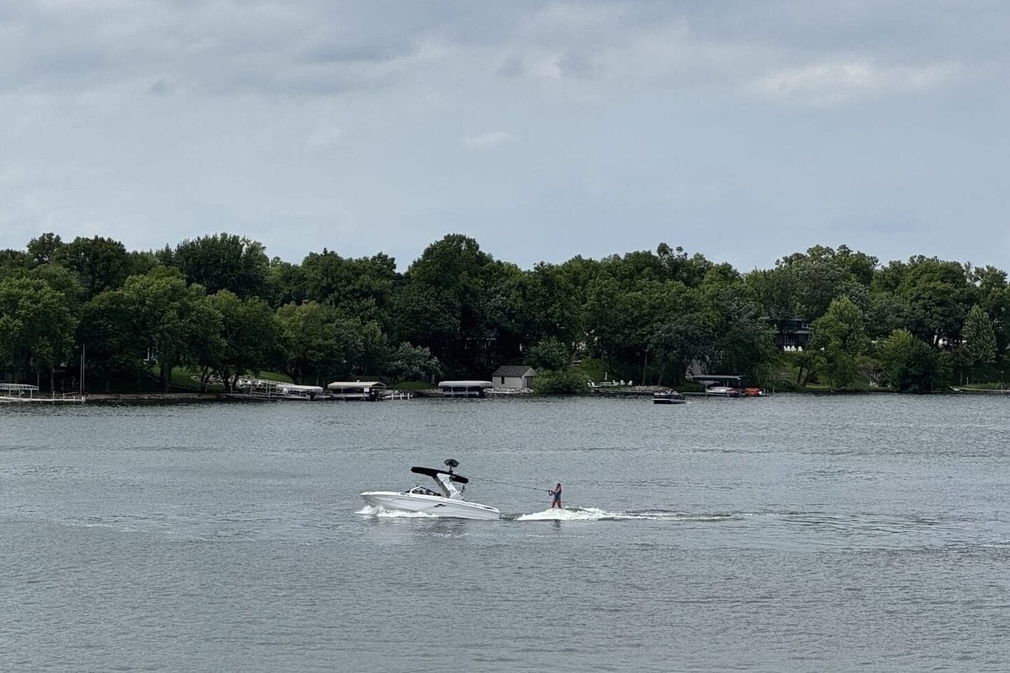 A person is wakeboarding behind a white boat on a lake, with another boat nearby and trees lining the shore under a cloudy sky.