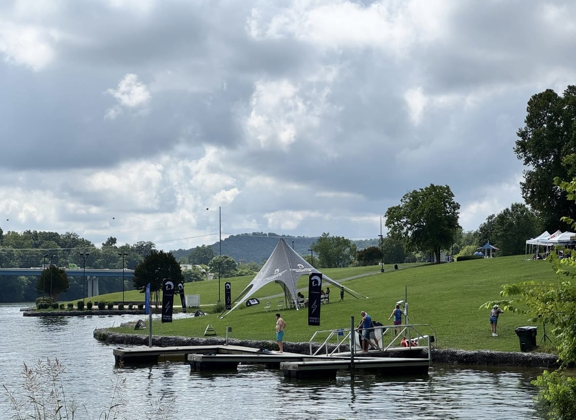 People stand on a dock and grassy shore beside a river. A white canopy tent and several banners are set up on the lawn. Trees and a bridge are visible in the background under a cloudy sky.