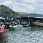 Boats docked at a marina with people nearby; American flags displayed, trees and a green bridge in the background under a partly cloudy sky.