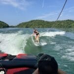 A person is wakesurfing behind a boat on a lake, while another person in a cap sits in the boat and watches. There are green hills and a blue sky in the background.