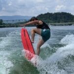 A person riding a bright red wakeboard performs a trick on the water, with a bridge and forested hills visible in the background.