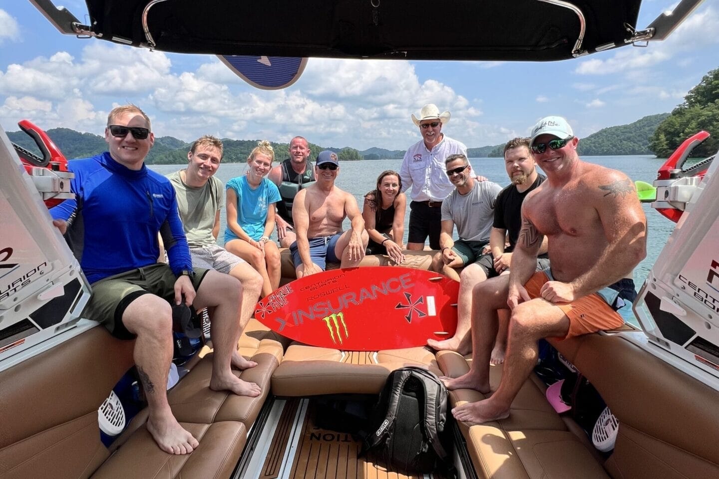 A group of people sit and pose for a photo on a Centurion Boat, some holding a red wakeboard during the Operation Wake Surf Honor Heroes Salute to Service Event on South Holston Lake, with trees and sky visible in the background.