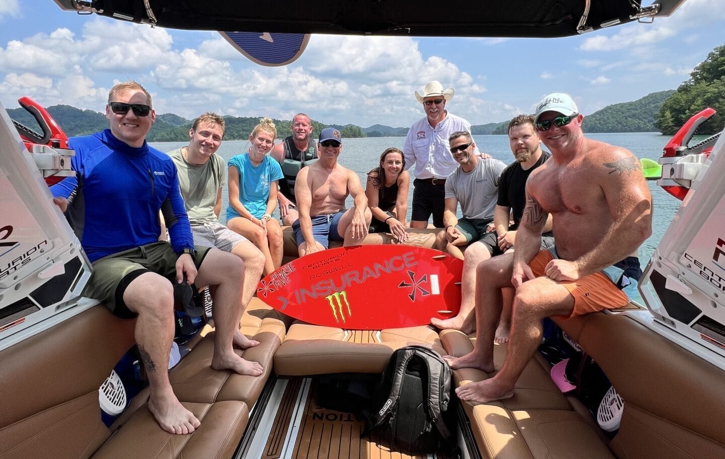 A group of eleven people pose together on a boat, holding a red wakeboard with 