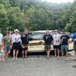 Eight men stand in front of a Tennessee State Trooper vehicle in a parking lot, holding beverages, with trees and hills in the background.