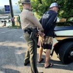 A police officer handcuffs a man in casual clothing next to a police vehicle in a parking lot near a lake.
