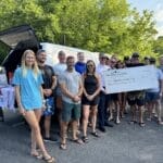 A group of people stand together outdoors near a van, holding a large ceremonial check made out to Operation Wake Surf. Towels and water coolers are visible.