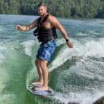 A man wearing a life vest and blue floral shorts is wakesurfing on a lake with green trees and a hillside in the background.